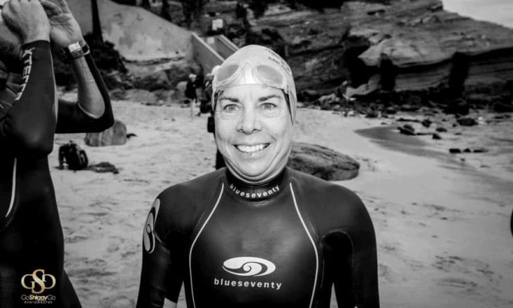 Rosalind Jarrett Sepulveda is photographed in black and white. She's wearing a wetsuit and swim cap and standing on a beach beside the water.